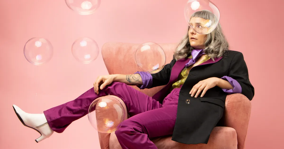 A woman with gray hair and glasses sitting in a pink armchair against a pink background. She is wearing a black blazer over a purple shirt and matching purple pants, with white high-heeled shoes. Bubbles float around her, adding a whimsical touch to the scene. She has a tattoo on her left arm and is looking off to the side with a thoughtful expression.